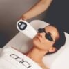 Woman lying on a spa bed wearing protective goggles while a clinician uses a handheld light/laser device near her face.