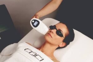 Woman lying on a spa bed wearing protective goggles while a clinician uses a handheld light/laser device near her face.
