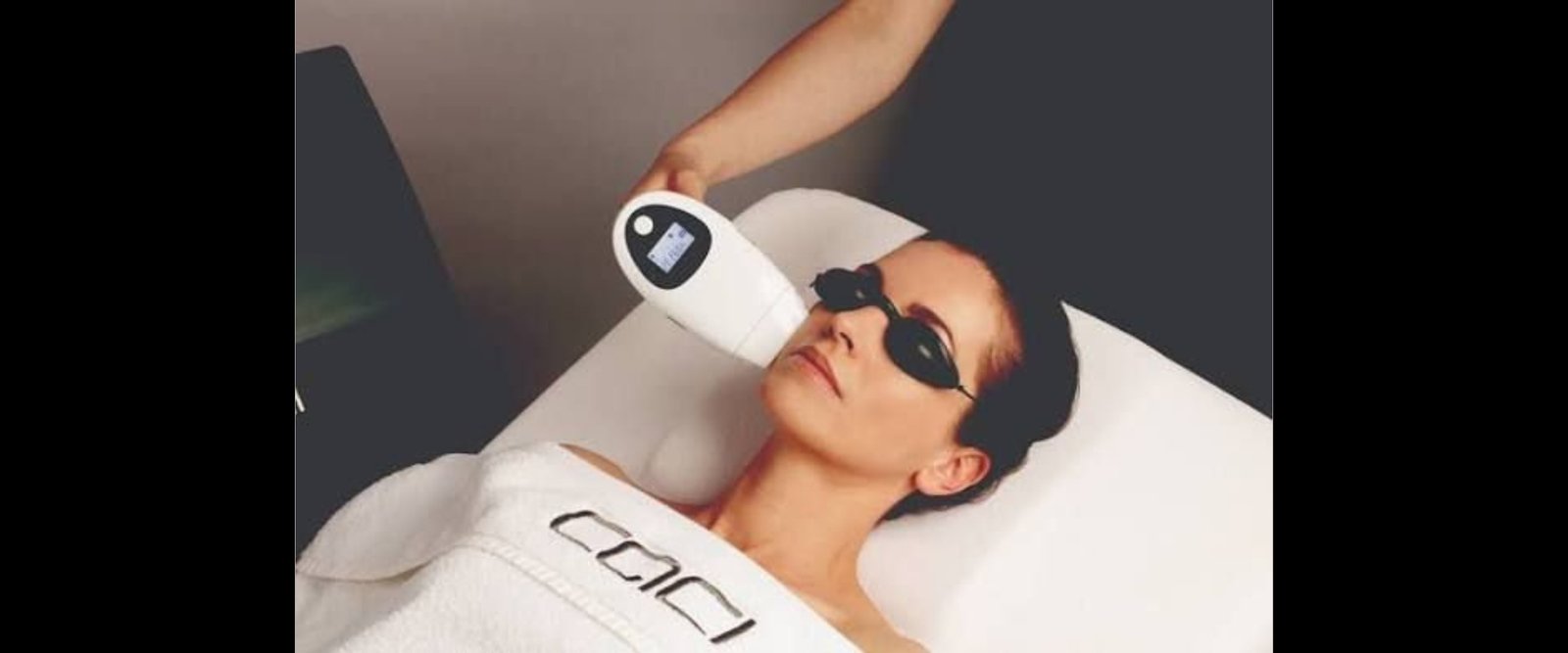 Person lying on a treatment bed with protective goggles as a clinician uses a handheld facial device near their face for light therapy.