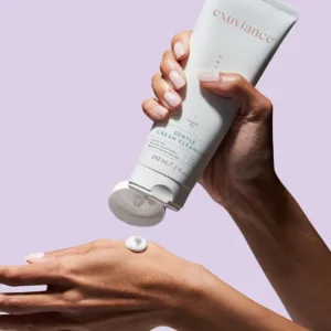 Hands dispense a white tube of Exuviance Gentle Cream Cleanser onto the back of the other hand against a lavender background.
