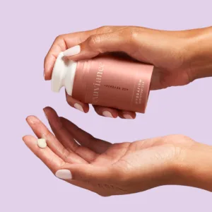 Hands hold a pink skincare bottle while a small amount of lotion sits on the other palm against a light purple background.