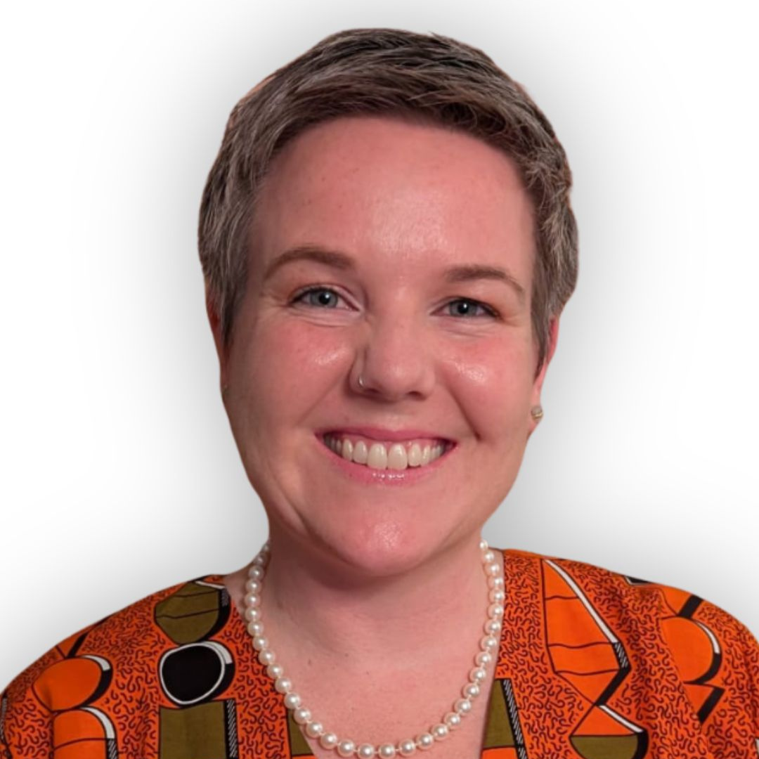 Smiling woman with short hair wearing an orange patterned top and a pearl necklace, portrait against a light background.