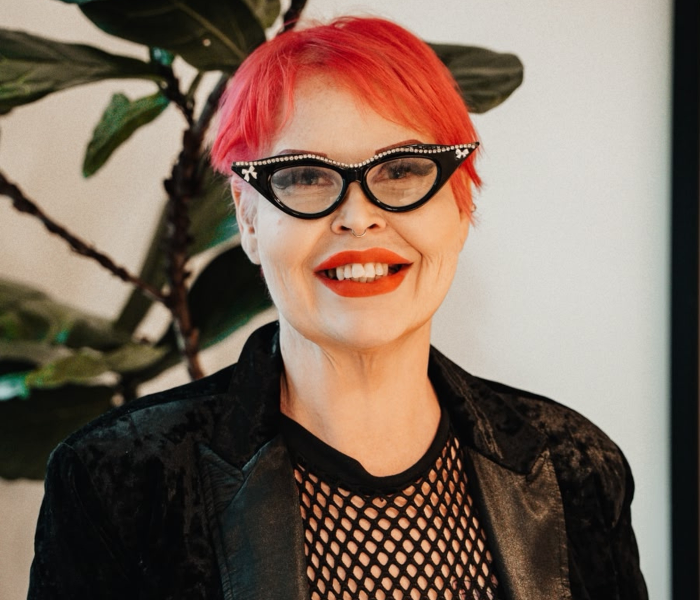 Portrait of a person with bright pink hair, rhinestone-edged cat-eye glasses, red lipstick, and a black blazer with a fishnet top, smiling indoors.