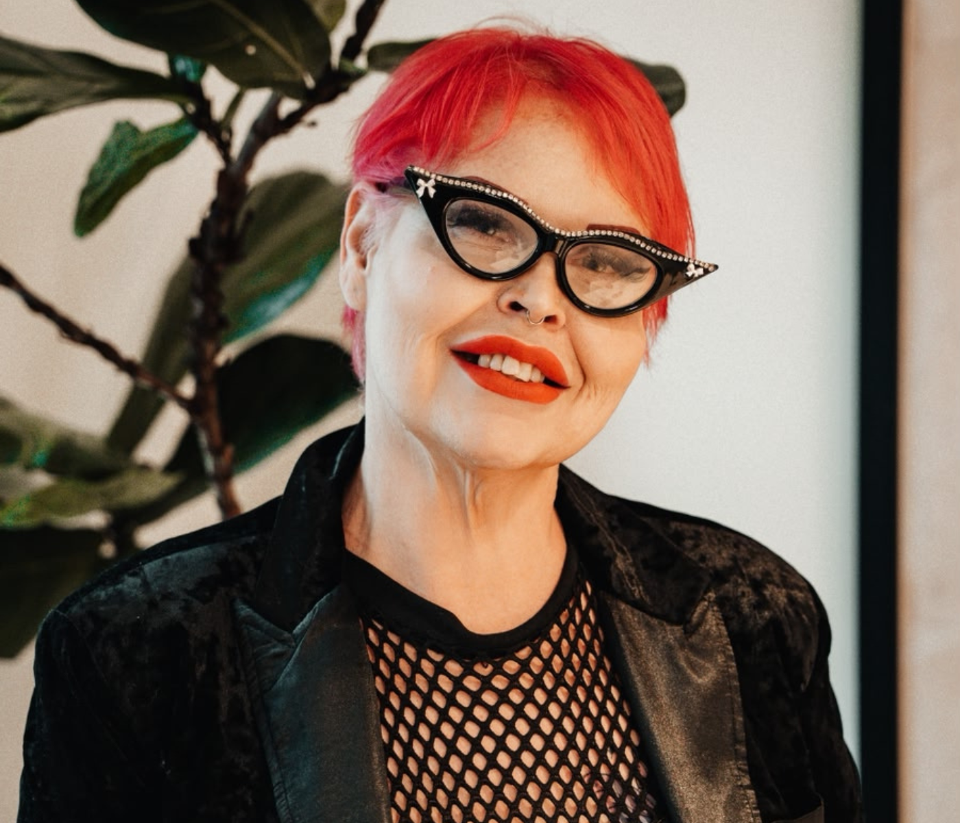 Portrait of a person with bright red hair, wearing rhinestone-edged cat-eye glasses, bold orange lipstick, and a nose ring, in a black blazer and fishnet top.