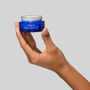 Blue cosmetic jar held between fingers showing open lid and cream; label reads Power Lift Advanced Ultra-Rich Moisturizer (in English and French).