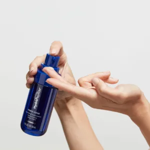 Close-up of hands twisting open the blue cap of a blue skincare serum bottle against a light background.