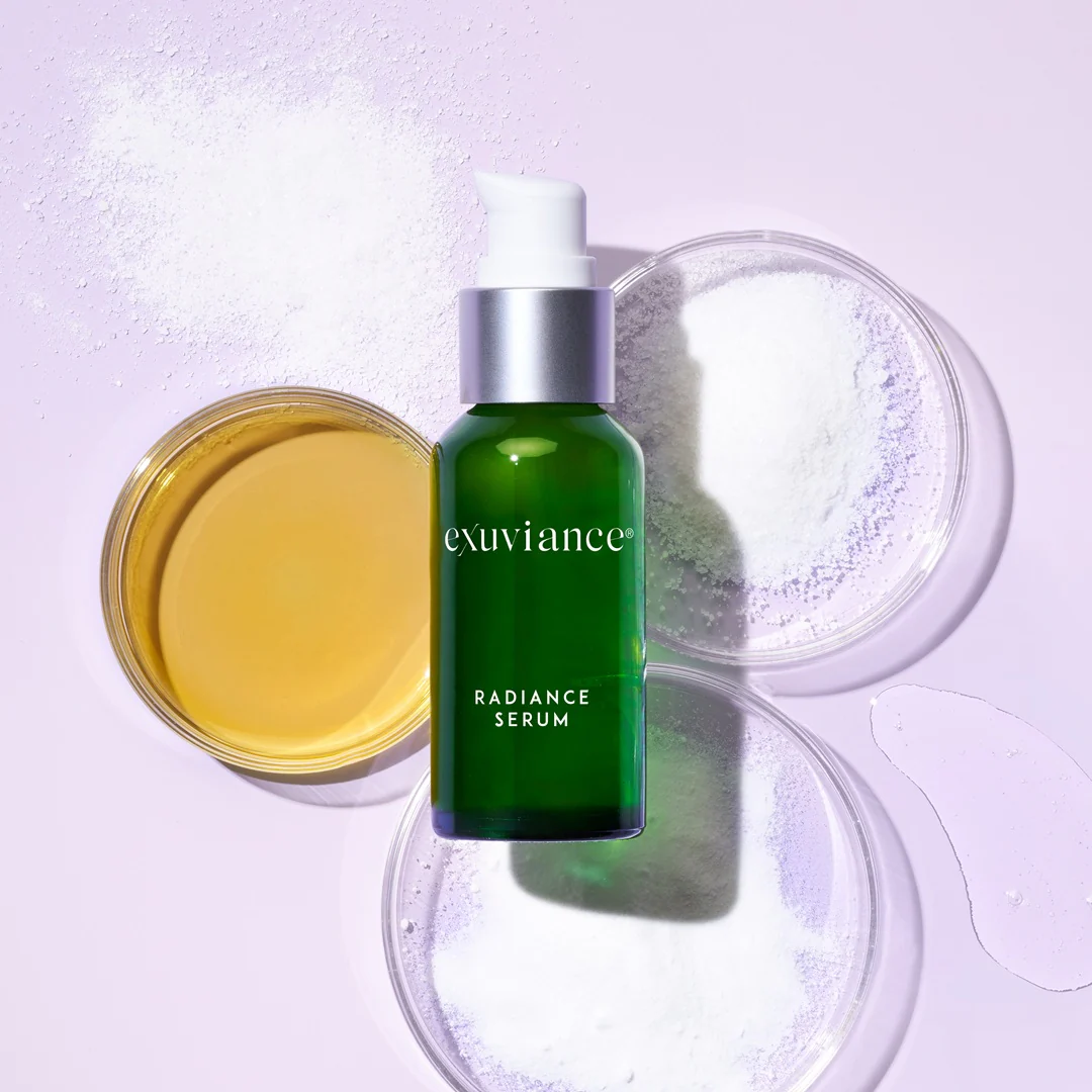 RadianceSerum_Ingredients - Dr Hans Clinics Exuviance Radiance Serum bottle with white pump, green glass, on a lavender background with scattered white powder.