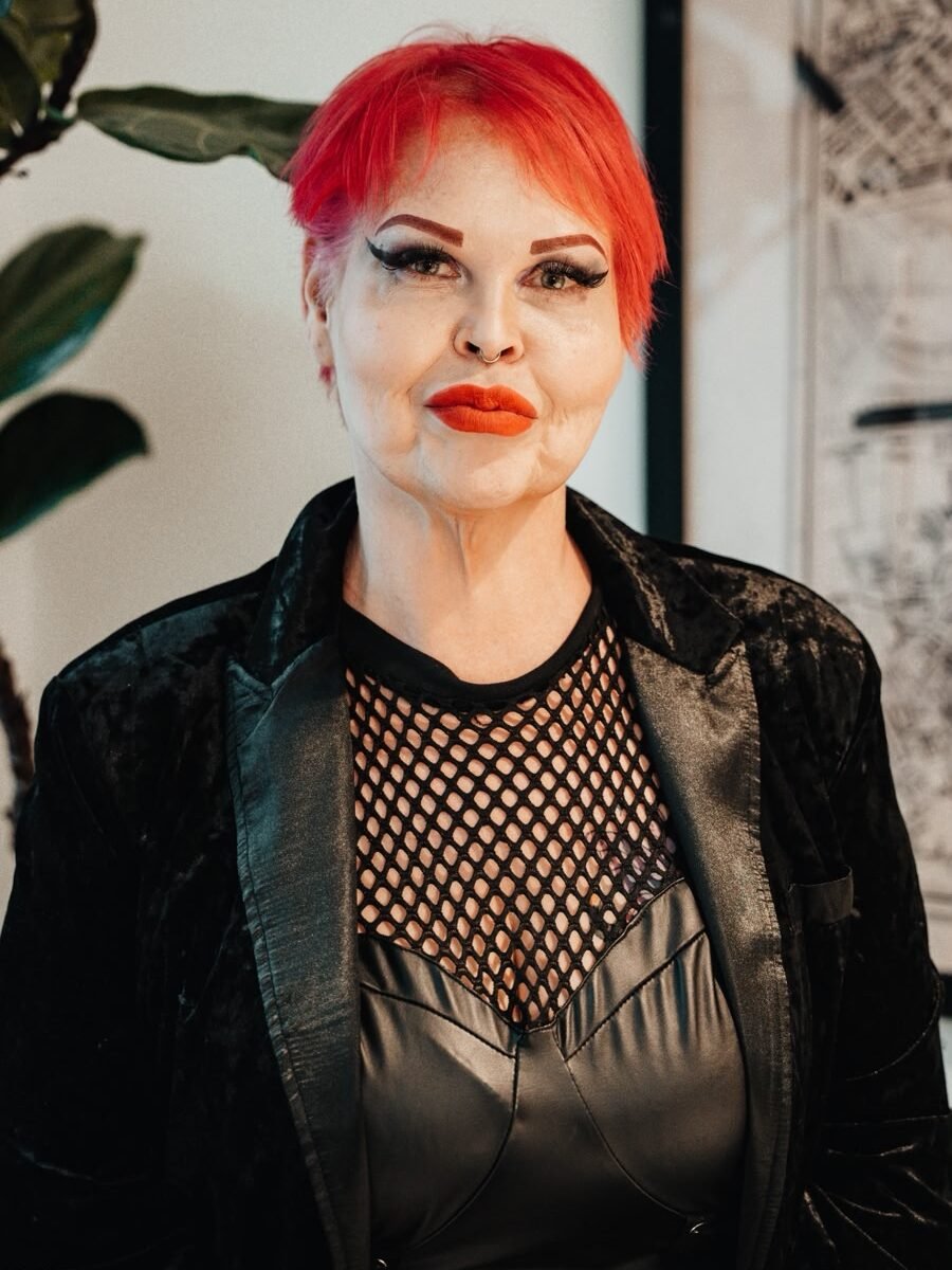Portrait of a person with short bright red hair, bold makeup, and a black fishnet top under a velvet blazer, indoors.