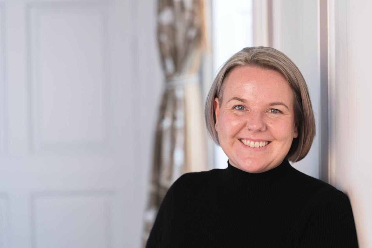 Smiling woman with short blonde hair in a black turtleneck, leaning against a wall indoors near a window.