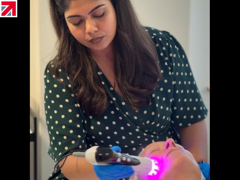 Clinician in a green polka dot dress wearing gloves uses a handheld laser device near a patient’s mouth during a cosmetic/dental procedure; patient lies back with glowing light from the device visible.