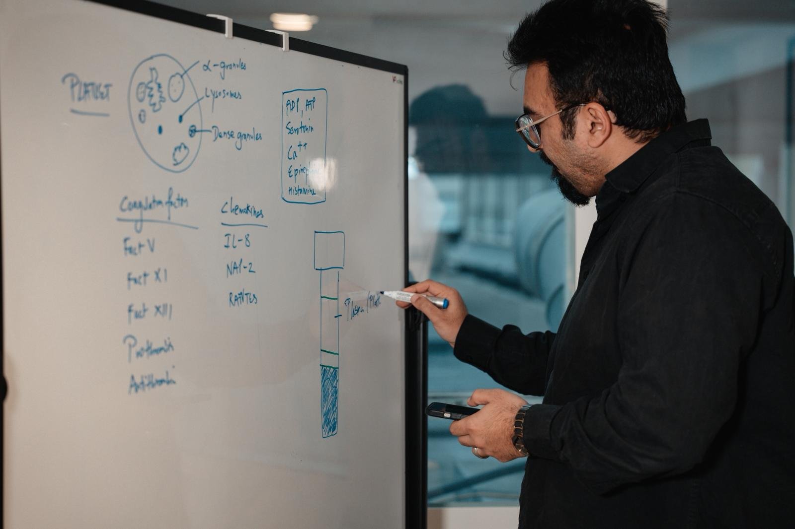 Man with glasses writes on a whiteboard with a marker while holding a smartphone.