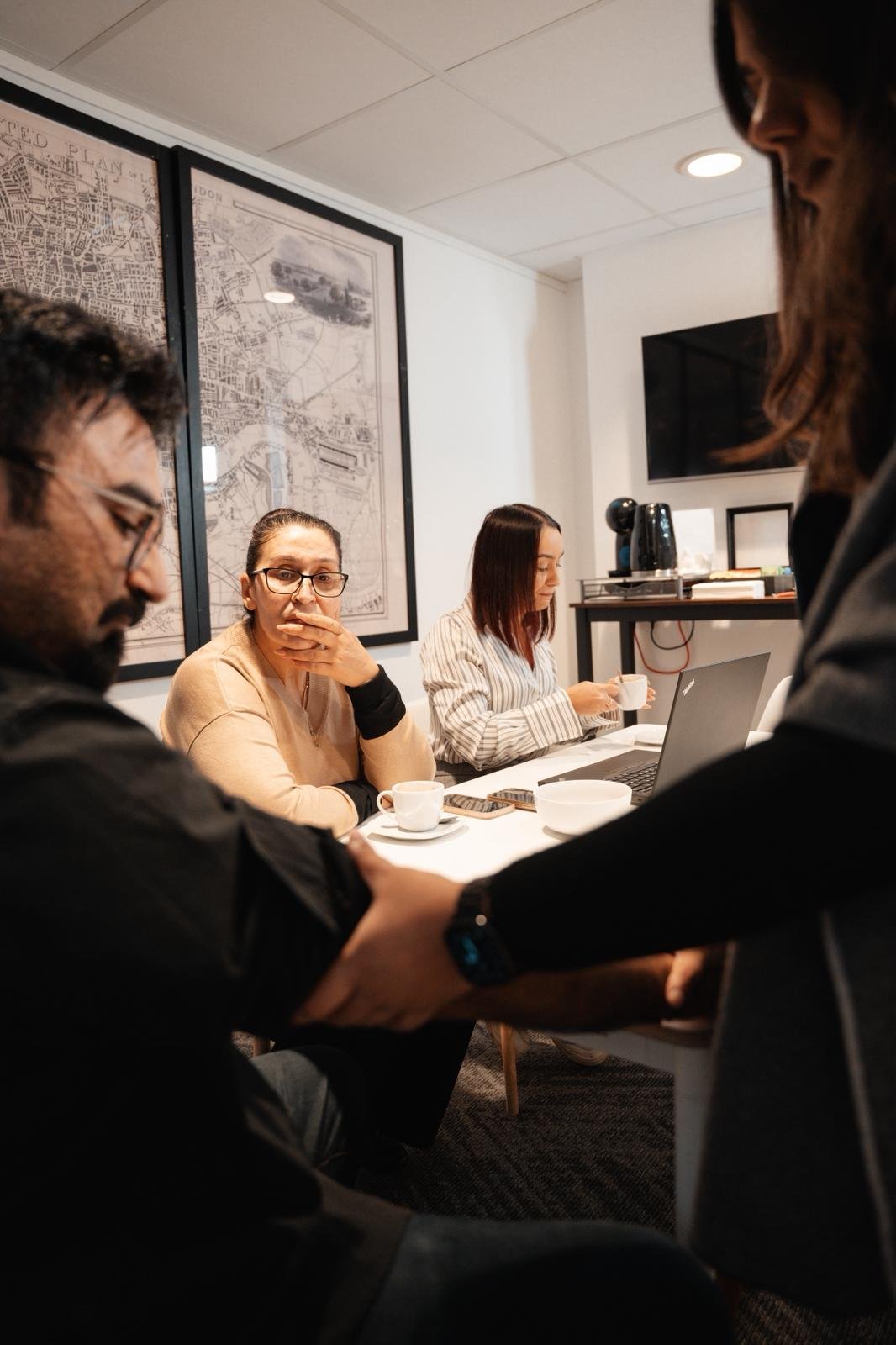 Team members in a meeting around a table with coffee cups; two people shake hands in the foreground while others work on laptops