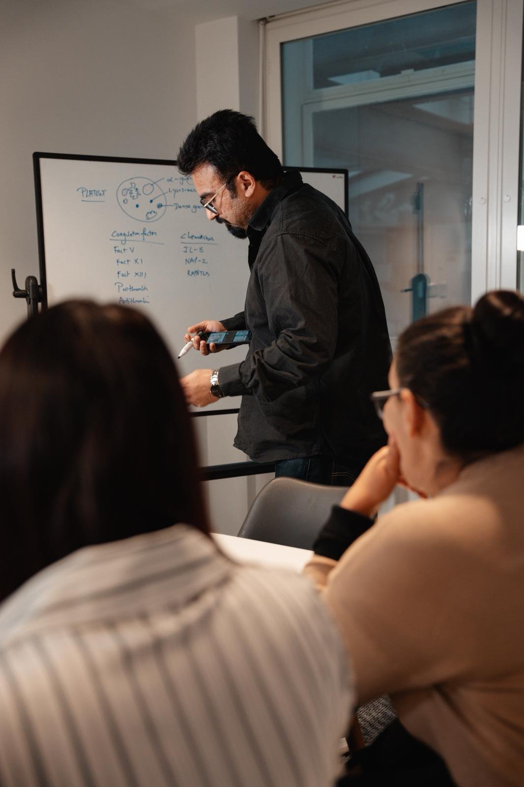 Instructor in a dark shirt stands by a whiteboard, holding a tablet as he explains to students seated in front of him.