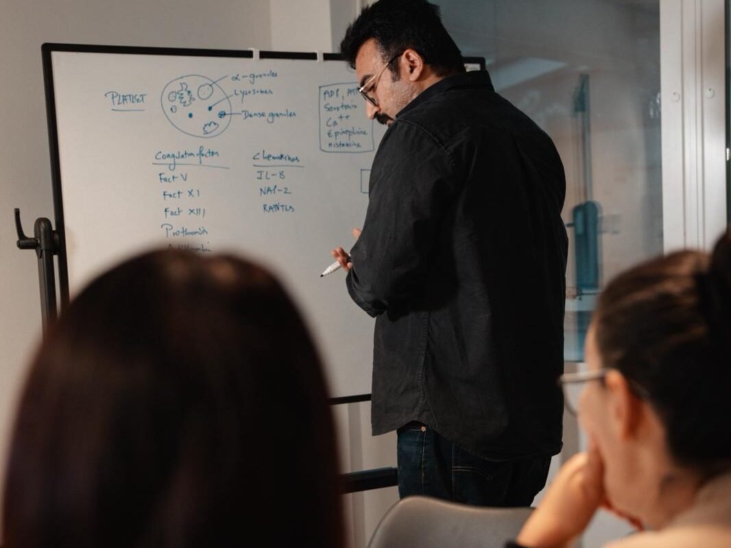 Instructor in a dark shirt writes on a whiteboard during a lecture, with students watching in the foreground.