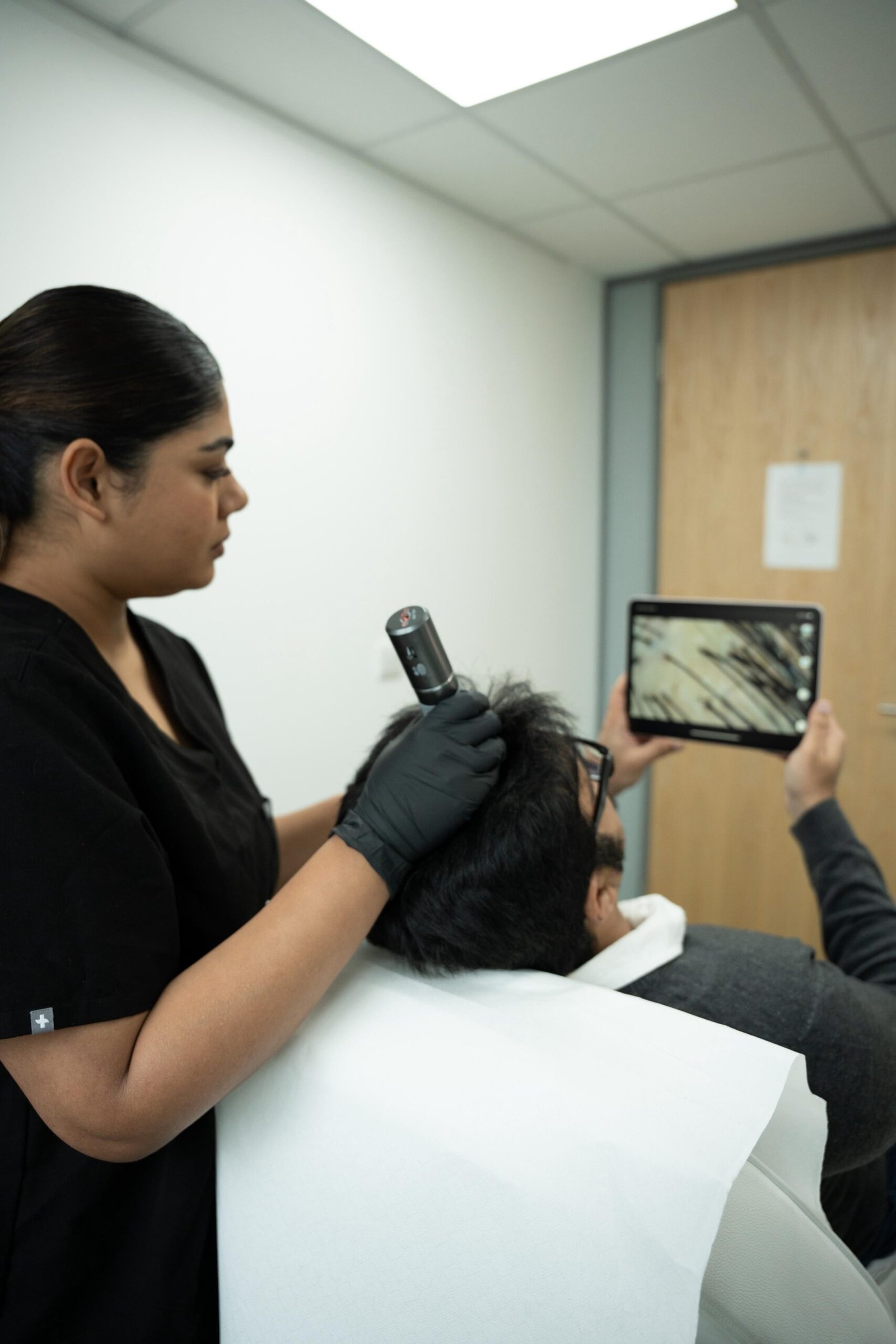 hair1 - Dr Hans Clinics - Dr Hans Clinics Hair stylist in black scrubs with gloves trims a client's hair, white cape draped over the client, while another person holds a tablet showing a photo.