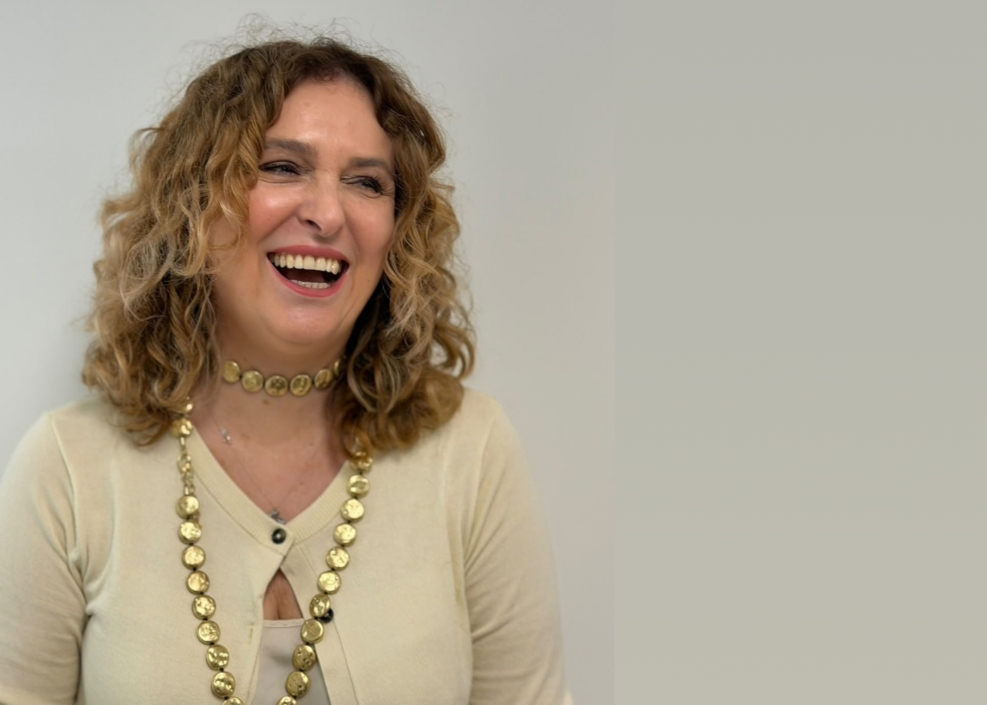 Smiling woman with curly light brown hair wearing a cream sweater and gold jewelry, looking to the side.