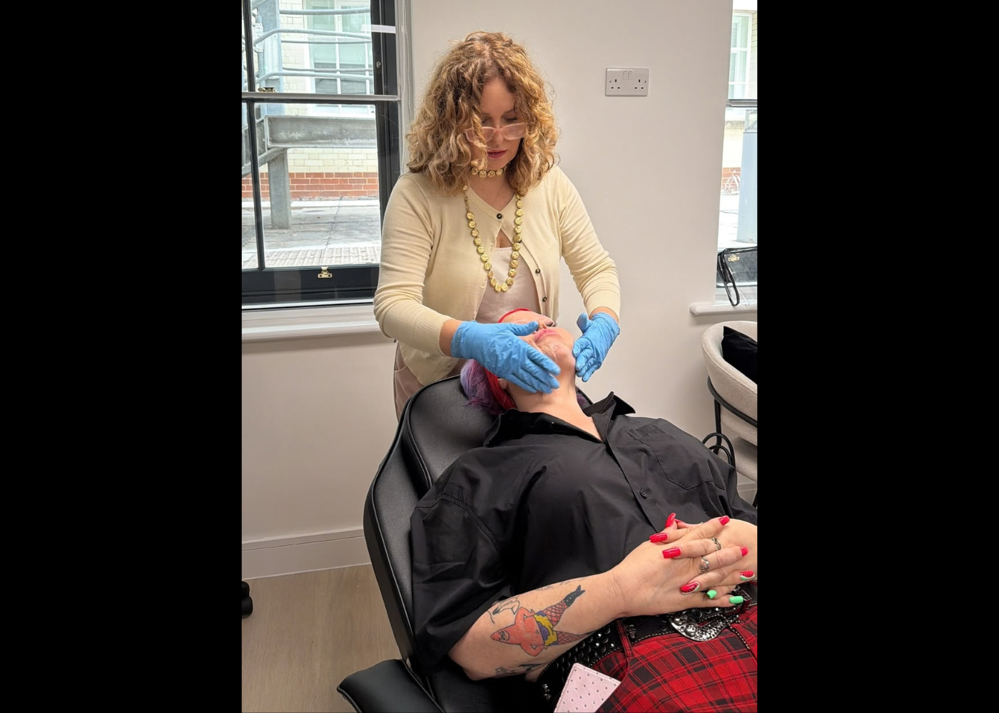Esthetician wearing a beige cardigan and blue gloves performs a facial on a client reclining in a salon chair by a window.