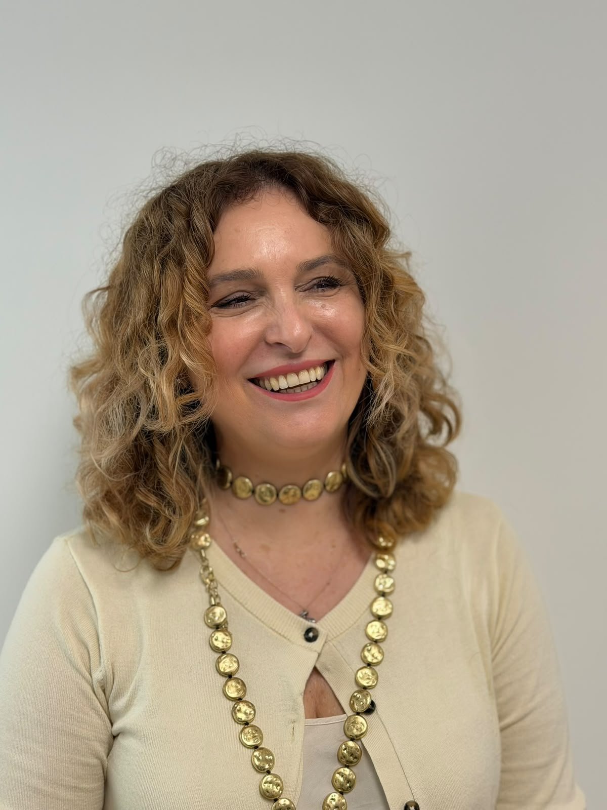 Smiling woman with curly light brown hair, wearing a beige cardigan and gold bead necklace.