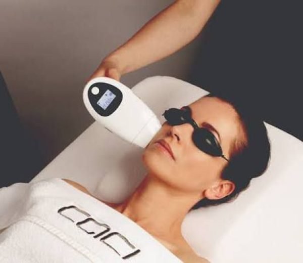 Woman lying on a spa bed wearing protective goggles while a clinician uses a handheld light/laser device near her face.
