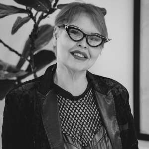 Smiling woman with short hair wearing rhinestone-edged cat-eye glasses and a dark blazer over a mesh top.